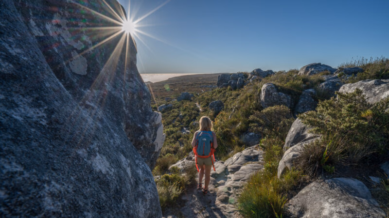 Hiker with a backpack on a rocky mountain trail with a bright sun star on luxury Cape Town holidays.