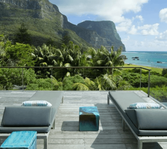 An outdoor sun terrace with two sun loungers on a wooden deck with a view of sea and mountains on Lord Howe Island