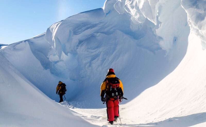 Two people in red and yellow jackets walk through a narrow snow gully towards a massive curved wall of ice.