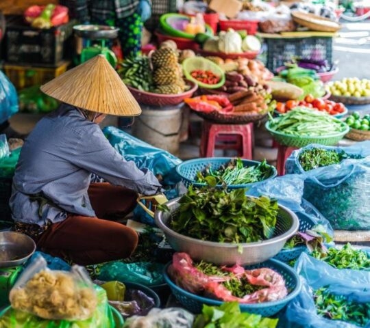 A person in a conical hat sorting fresh vegetables at a colorful open-air market stall filled with produce.