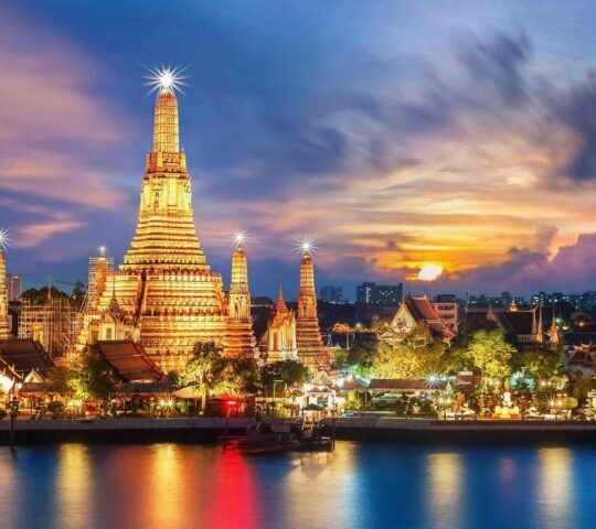 The grand Wat Arun temple complex in Bangkok glows with golden lights at sunset by the river.