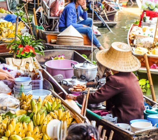 A person in a wooden boat sells yellow bananas and cooked food at a crowded Thai floating market.