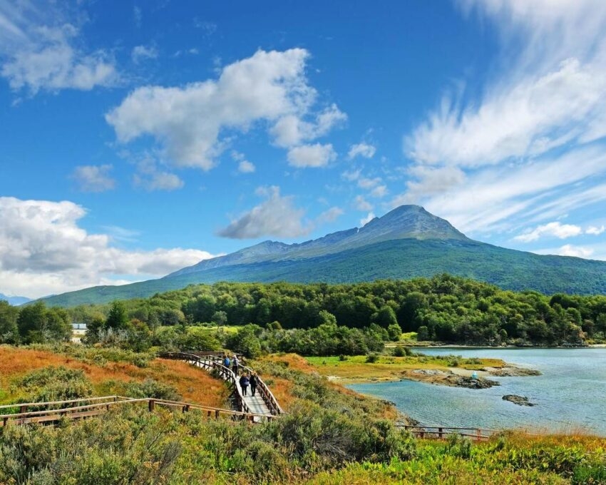 Panoramic view of Tierra del Fuego National Park, near Ushuaia , Argentina, showing people walking on a wooden bridge towards a volcano surrounded by green vegetation and water, against a blue sky covered by a few white clouds.. Panoramic view of Tierra del Fuego National Park, showing a volcano surrounded by green vegetation and water, against a blue sky. ushuaia, tierra, fuego, national, park, nature, green, water, blue, volcano, mountain, bridge, wood, people, brown, clouds, white, landscape, argentina