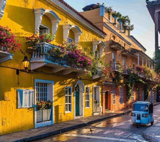 Sunlit street in Cartagena, featuring vibrant yellow walls adorned with colorful flowers, and a blue tuk-tuk parked nearby.