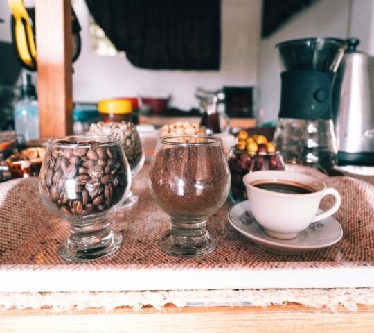 A wooden tray holds three glass containers with coffee beans, ground coffee, and a white cup of coffee, surrounded by various snacks.