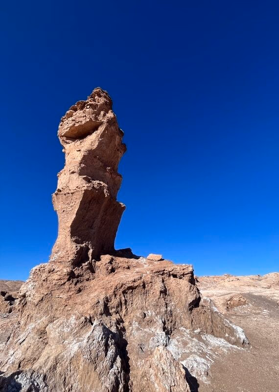 A tall, rugged rock formation stands against a clear blue sky, showcasing the arid landscape of a desert region.
