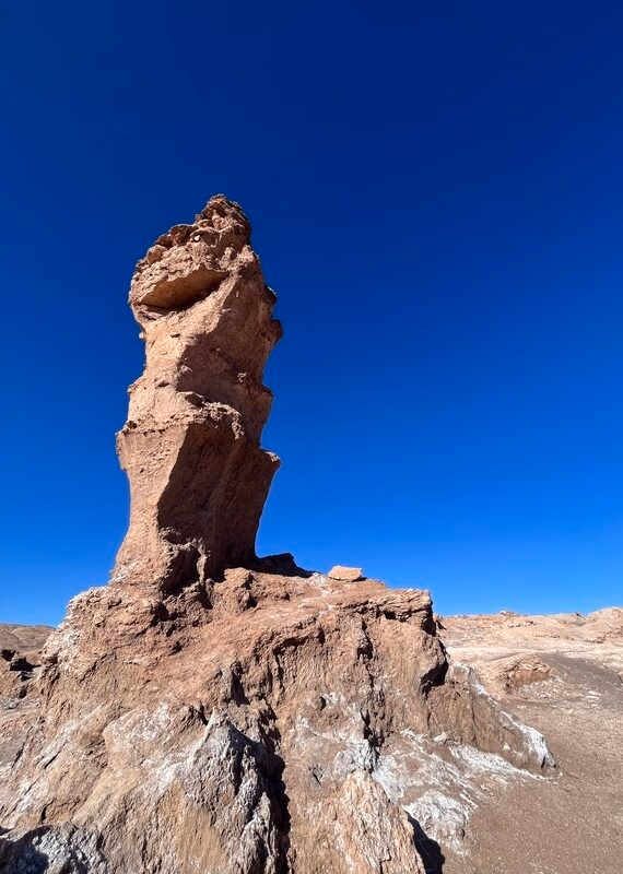 A tall, rugged rock formation stands against a clear blue sky, showcasing the arid landscape of a desert region.