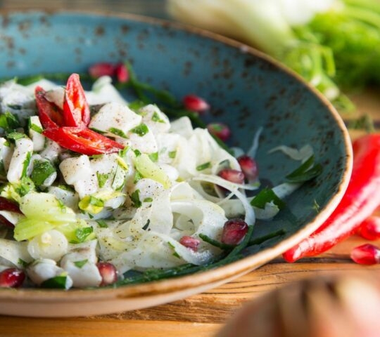 A vibrant salad featuring thinly sliced vegetables, pomegranate seeds, and garnished with red chili, served in a rustic bowl.