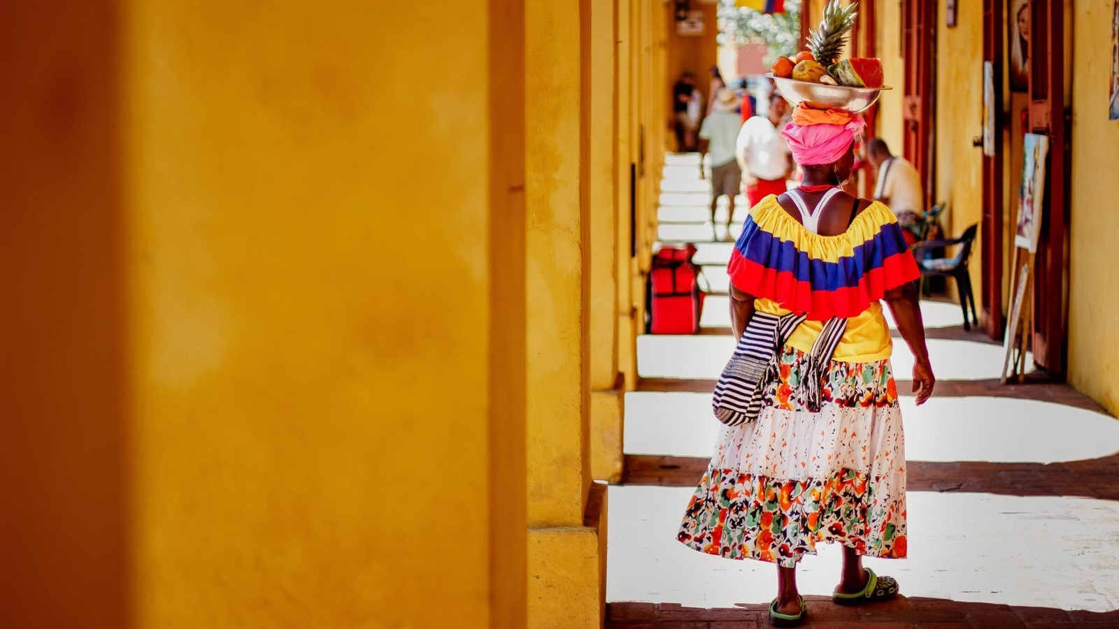 A woman in colorful traditional attire walks through a sunlit yellow corridor, balancing a bowl of fruits on her head.
