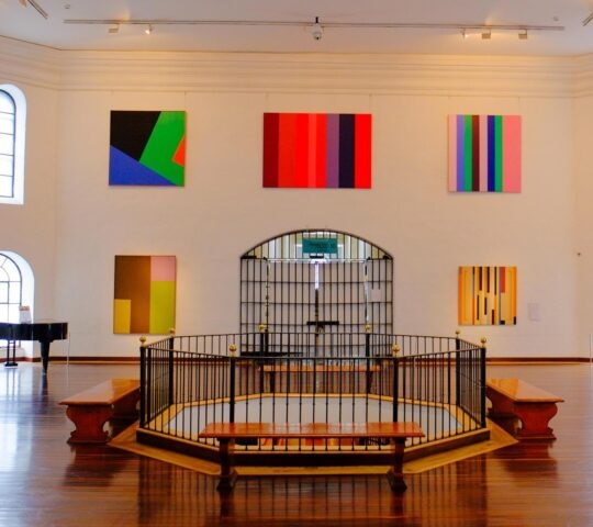 A modern art gallery featuring colorful abstract paintings on white walls, with hardwood floors and a grand piano in the corner.
