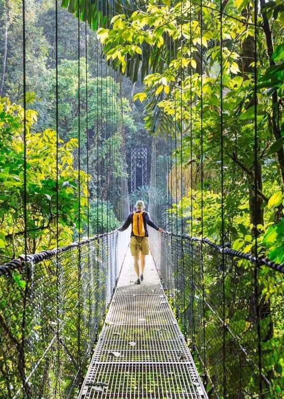 A hiker with a yellow backpack walking across a narrow suspension bridge in a lush green jungle.