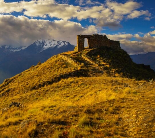 Ancient stone gateway on a mountain ridge at sunset with snow-capped peaks in the distance.