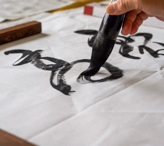 A hand holding a calligraphy brush painting black ink characters on white paper.