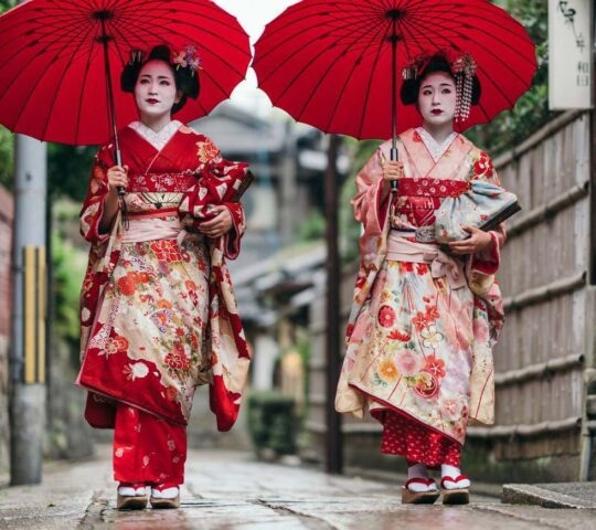 Two individuals in traditional Japanese kimonos holding red umbrellas on a street.