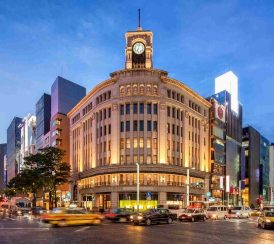Illuminated city corner with a classic building and clock tower at twilight, busy street with motion-blurred vehicles.