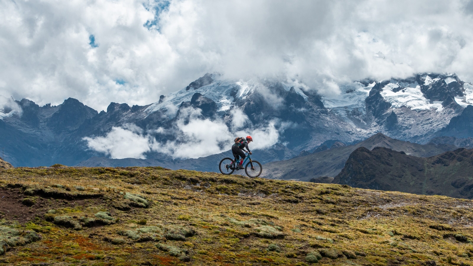 Cyclist mountain biking against a backdrop of clouds and snowy peaks.