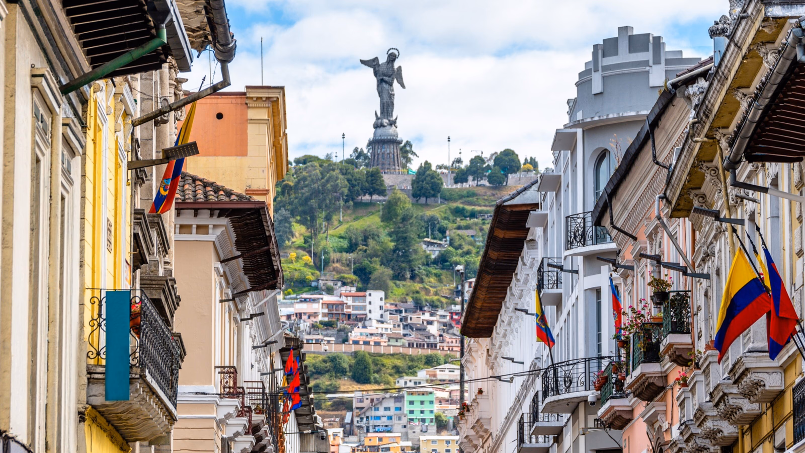A historic street in Quito with buildings flanking both sides and the El Panecillo statue in the background.