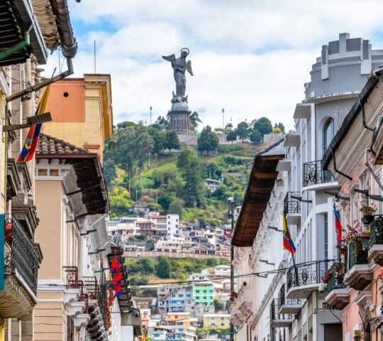 A historic street in Quito with buildings flanking both sides and the El Panecillo statue in the background.