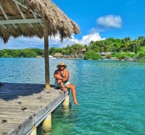 Person sitting on a tropical wooden dock with thatched roof, clear blue water, and lush greenery.