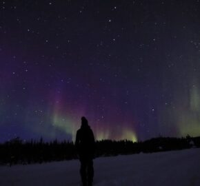 Person standing on snow under a starry sky with colorful northern lights.