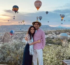 Couple embracing in a field with hot air balloons in the sky.