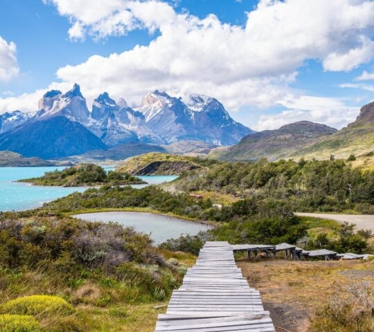 A wooden walkway leading towards turquoise lakes and rugged mountains under a blue sky.