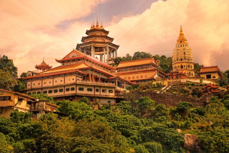 Traditional Asian pagoda and temple complex on a hillside with lush greenery under a warm, cloudy sky.