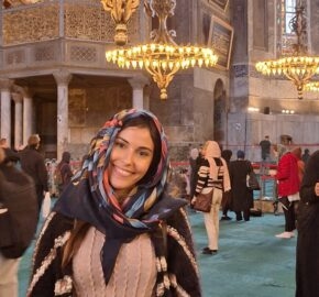 An individual in a patterned scarf inside an ornate room with a chandelier and visitors.