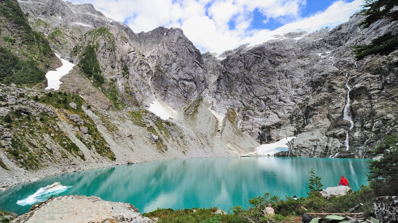 Person in red overlooking a turquoise mountain lake with icebergs and rugged cliffs.