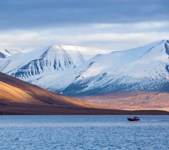 A red boat on a tranquil lake against a backdrop of snowy mountains under a soft blue sky.