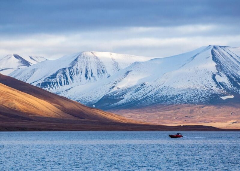 A red boat on a tranquil lake against a backdrop of snowy mountains under a soft blue sky.