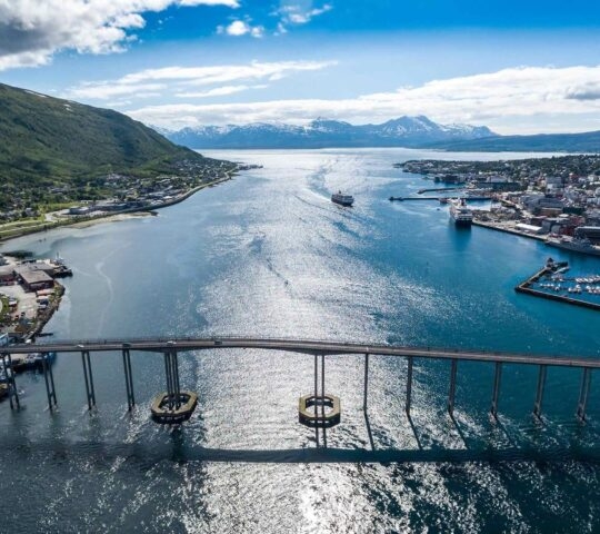 Aerial view of a coastal city with a bridge over a bay, surrounded by mountains, under a clear blue sky.