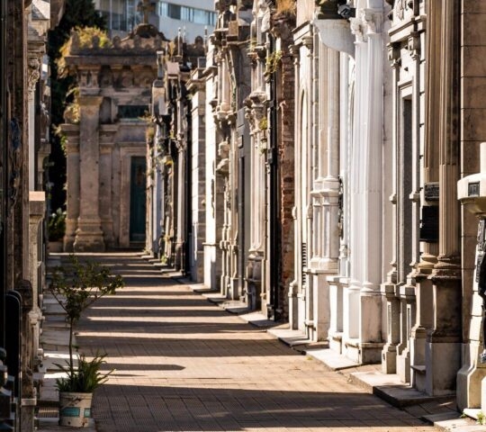Shadowed walkway with ornate mausoleums in a sunny cemetery.