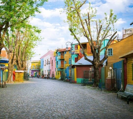 Cobbled street lined with colorful buildings and leafy trees, benches on the side, under a clear sky.