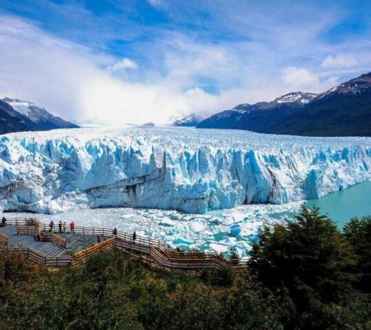 Observation deck with visitors overlooking a vast, towering glacier with a backdrop of mountains and clear skies.