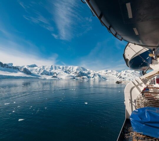 View from a ship's deck overlooking icy waters and snow-capped mountains under blue skies.