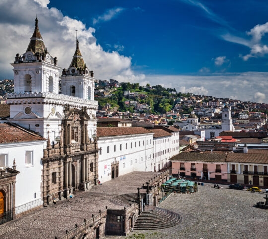 Historic white church and plaza with a backdrop of terracotta-roofed houses and hills against a cloudy blue sky.