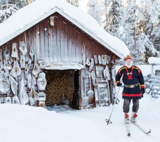 A Sami man stands on skis outside a snow covered lodge in Finnish Lapland