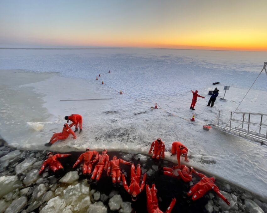 People in red suits on an ice-covered landscape with a sunset in the background.