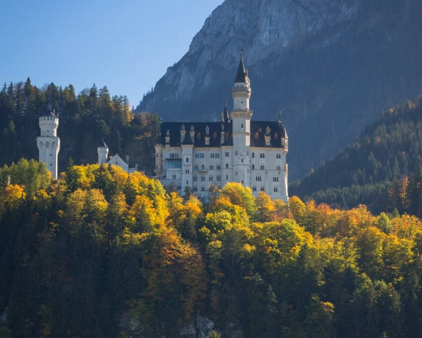 Neuschwanstein Castle atop a hill surrounded by autumn-colored trees with a clear blue sky.