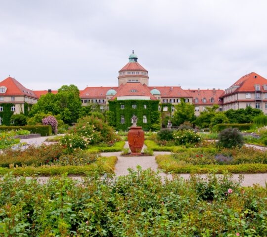 Formal garden with flowerbeds and sculptures in front of a historic building with a red roof.