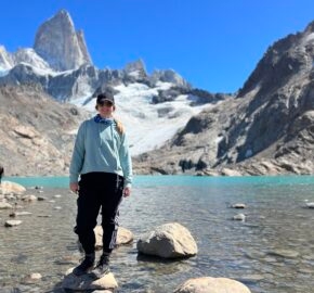 Person standing on a rock by a mountain lake with a glacier and peaks in the background.