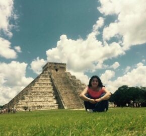 Person sitting on grass in front of El Castillo pyramid in Chichen Itza, Mexico, with blue sky and clouds.