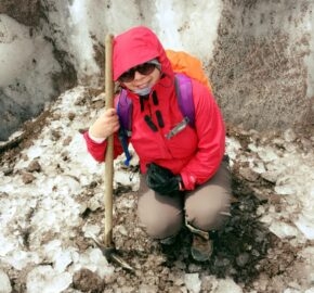 A hiker in a red jacket with a backpack and trekking pole on snowy terrain.