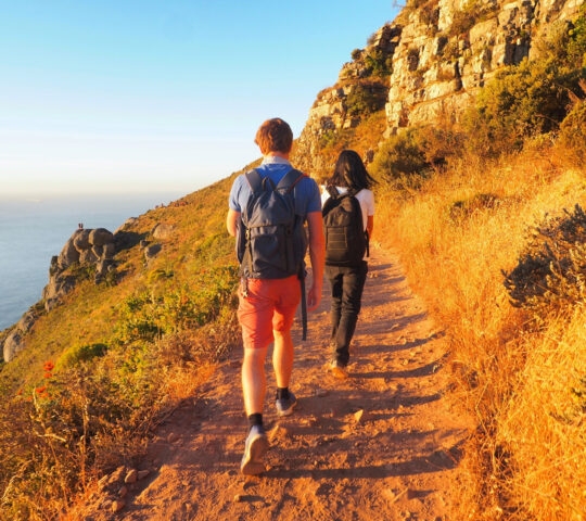 Two hikers with backpacks walking on a mountain trail at sunset.