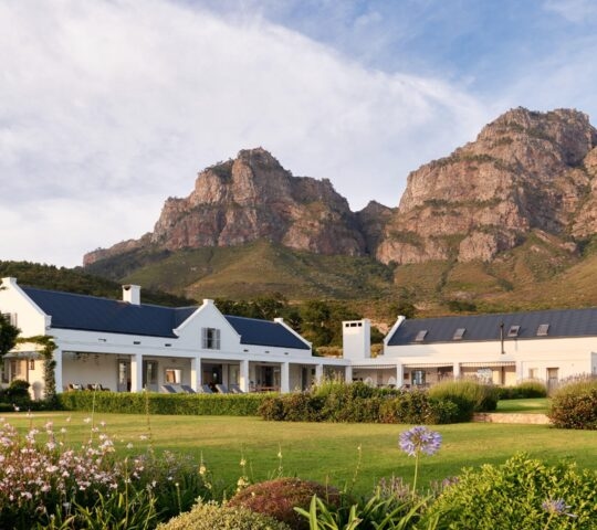 White house with blue roof in front of a mountain and garden.
