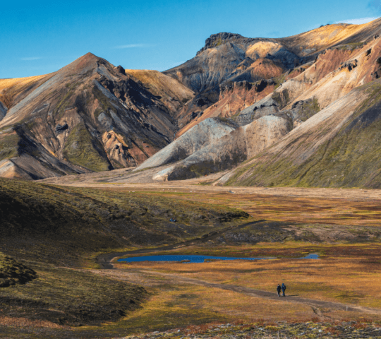 Two hikers walk through a colorful mountainous landscape with a blue pond under a clear sky.