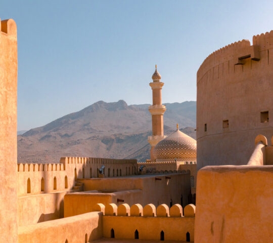 An ancient fortress with domes and a minaret set against a mountain backdrop.