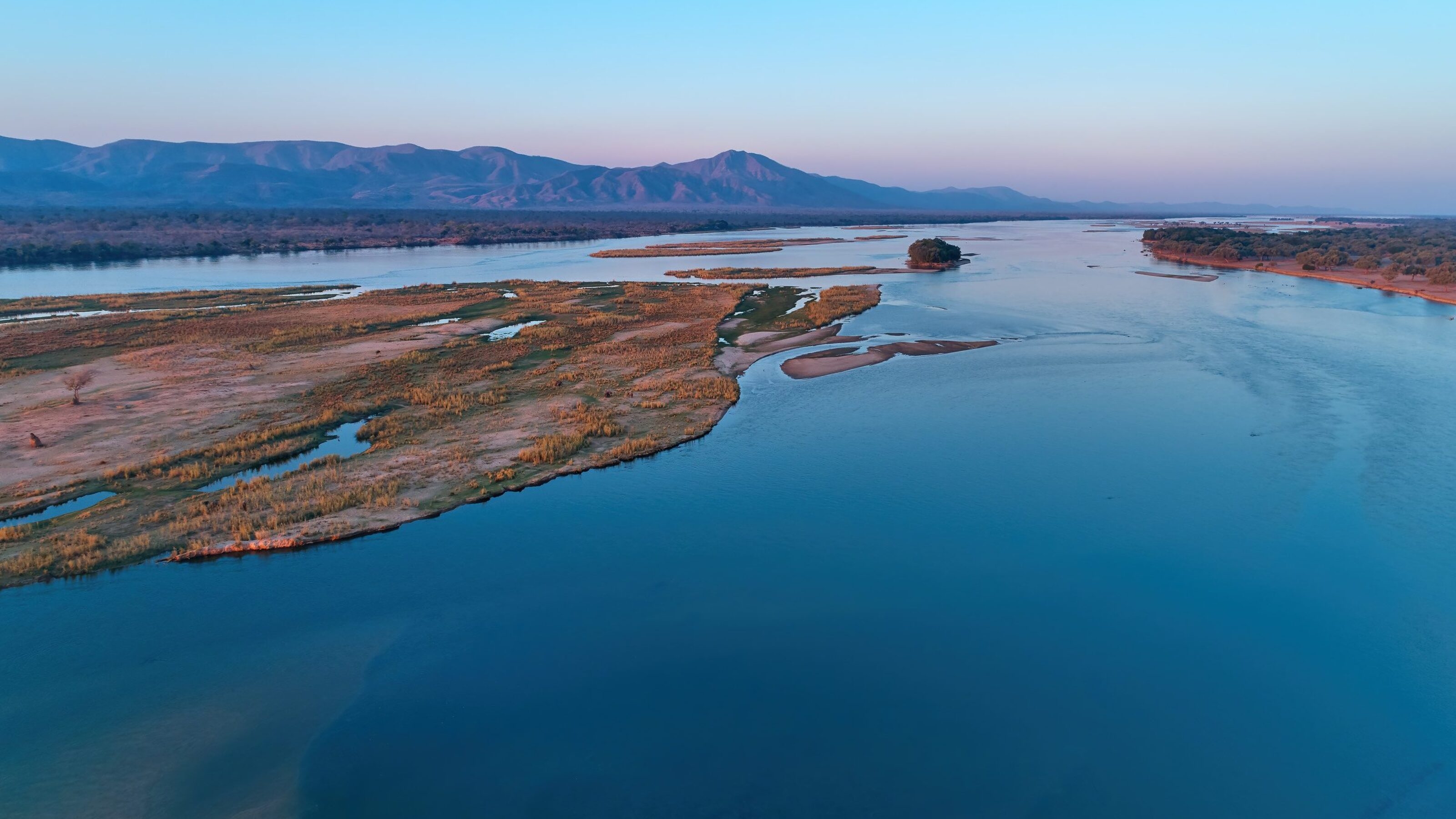 Aerial view of a serene river at dusk with surrounding wetlands and distant mountains.