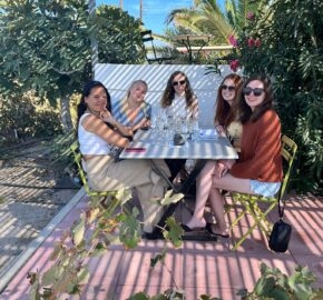 Group of people sitting around a table with glasses, surrounded by greenery under sunlight.